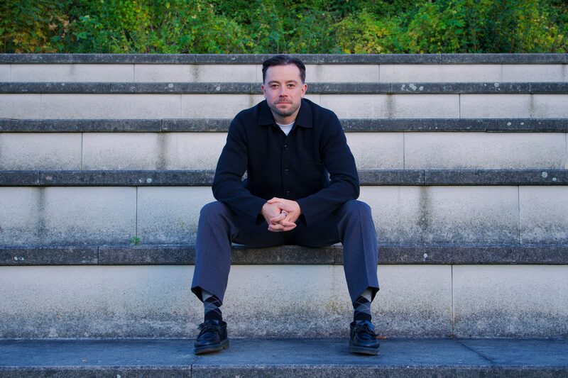 Theon Bower sitting on stone steps in Berlin, wearing a dark overshirt, looking at the camera