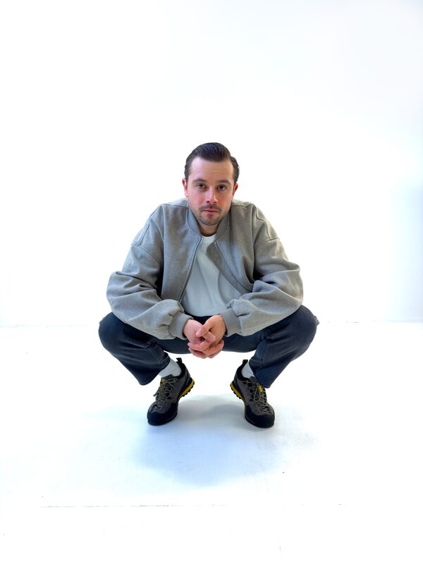 Theon Bower crouching in a white studio, wearing a grey bomber jacket